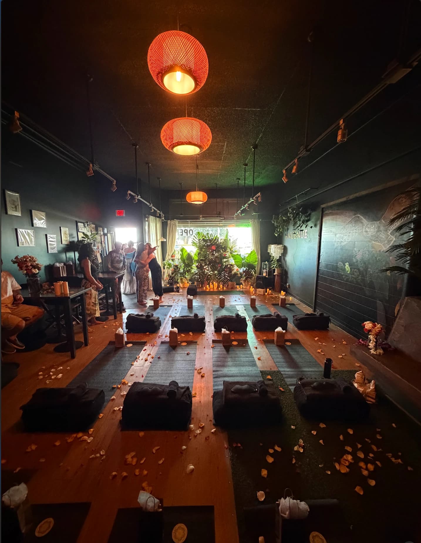 The Ānuenue lounge set for a candlelit sound bath: rows of yoga mats and dark bolsters on the honey wood floor, flower petals scattered between them, amber woven lanterns overhead, candles by the plant-filled front window, and the owl mural on the right wall